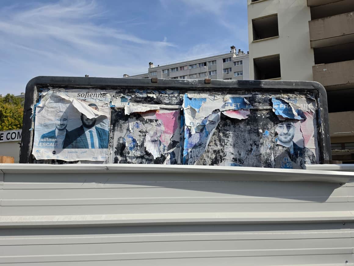 Panneau d’affichage couvert de portraits électoraux lacérés devant une palissade claire et des immeubles collectifs