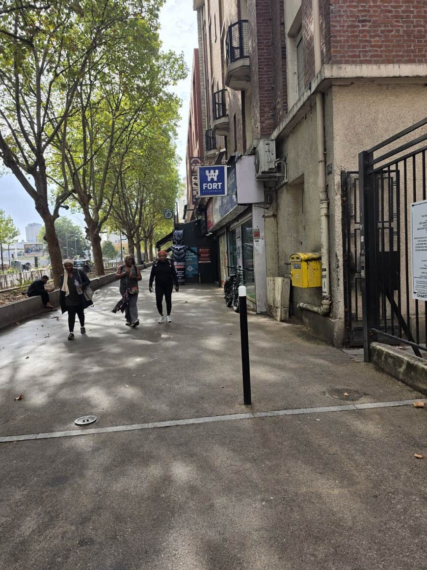 Trois femmes marchant sur un large trottoir arboré devant des commerces et une boîte aux lettres jaune