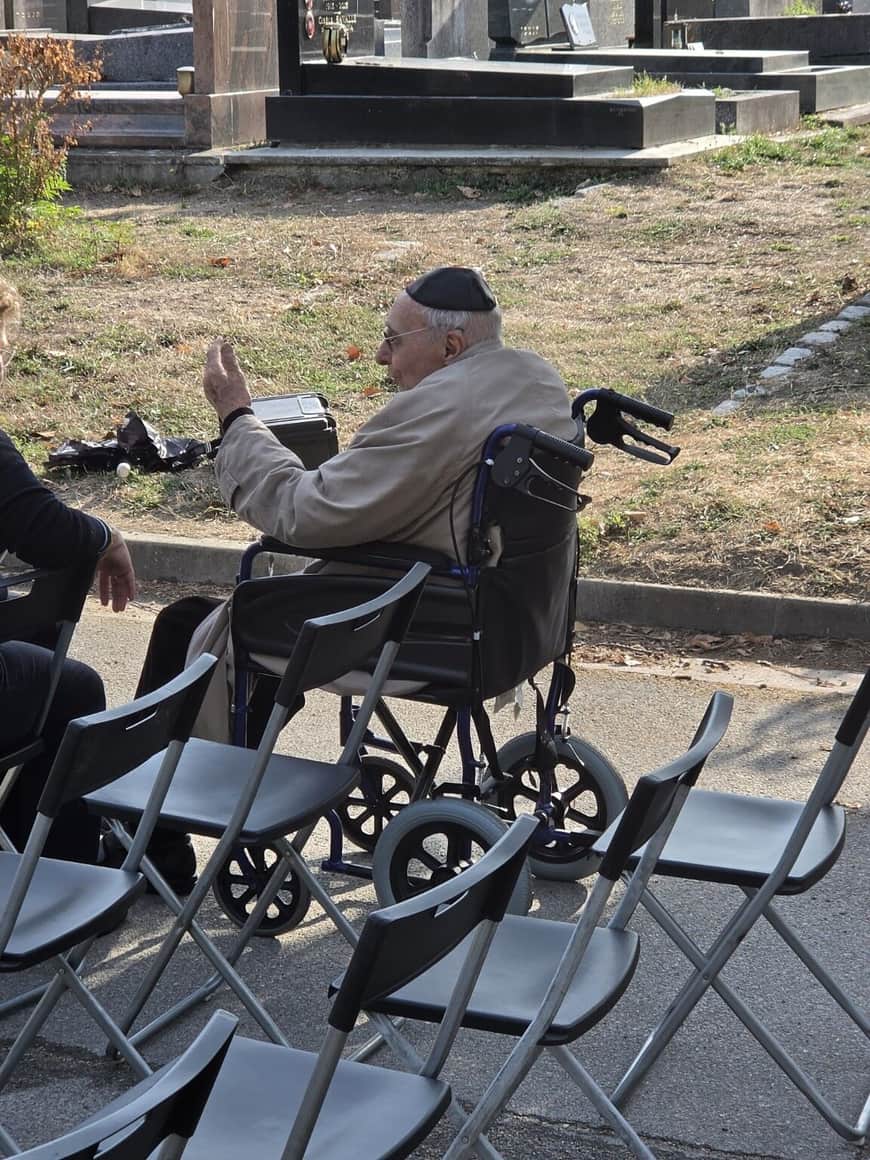 Homme âgé coiffé d’une kippa noire, assis dans un fauteuil roulant parmi des chaises pliantes et levant la main