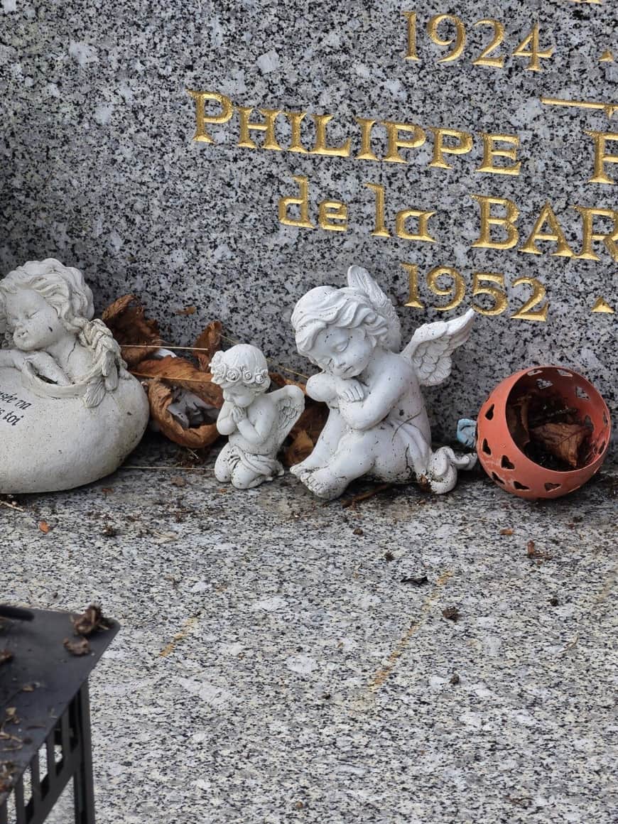 Trois figurines d’anges blancs disposées au pied d’une inscription funéraire dorée, parmi des feuilles mortes