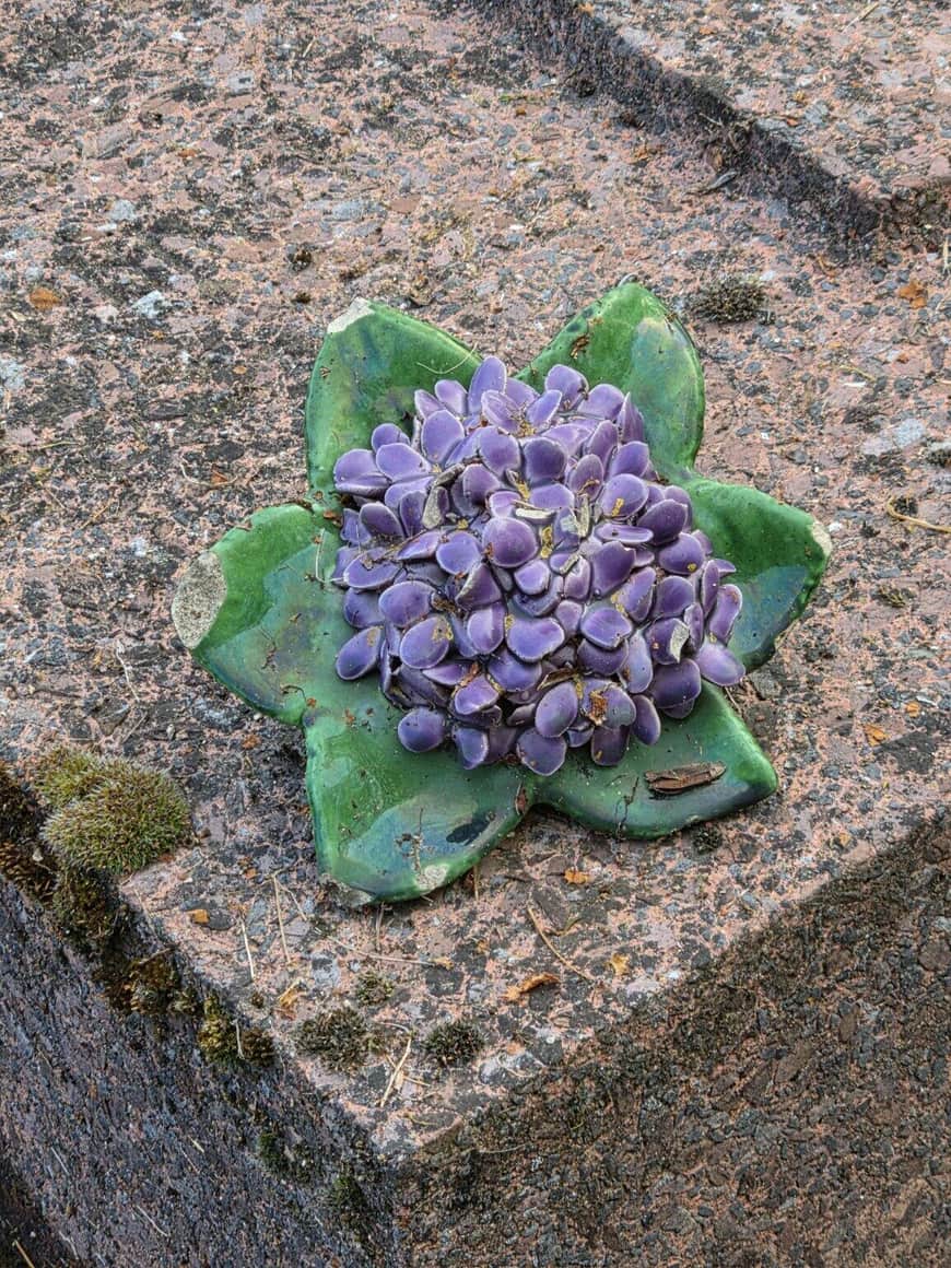 Ornement funéraire en céramique représentant un hortensia violet aux feuilles vertes, posé sur une pierre altérée