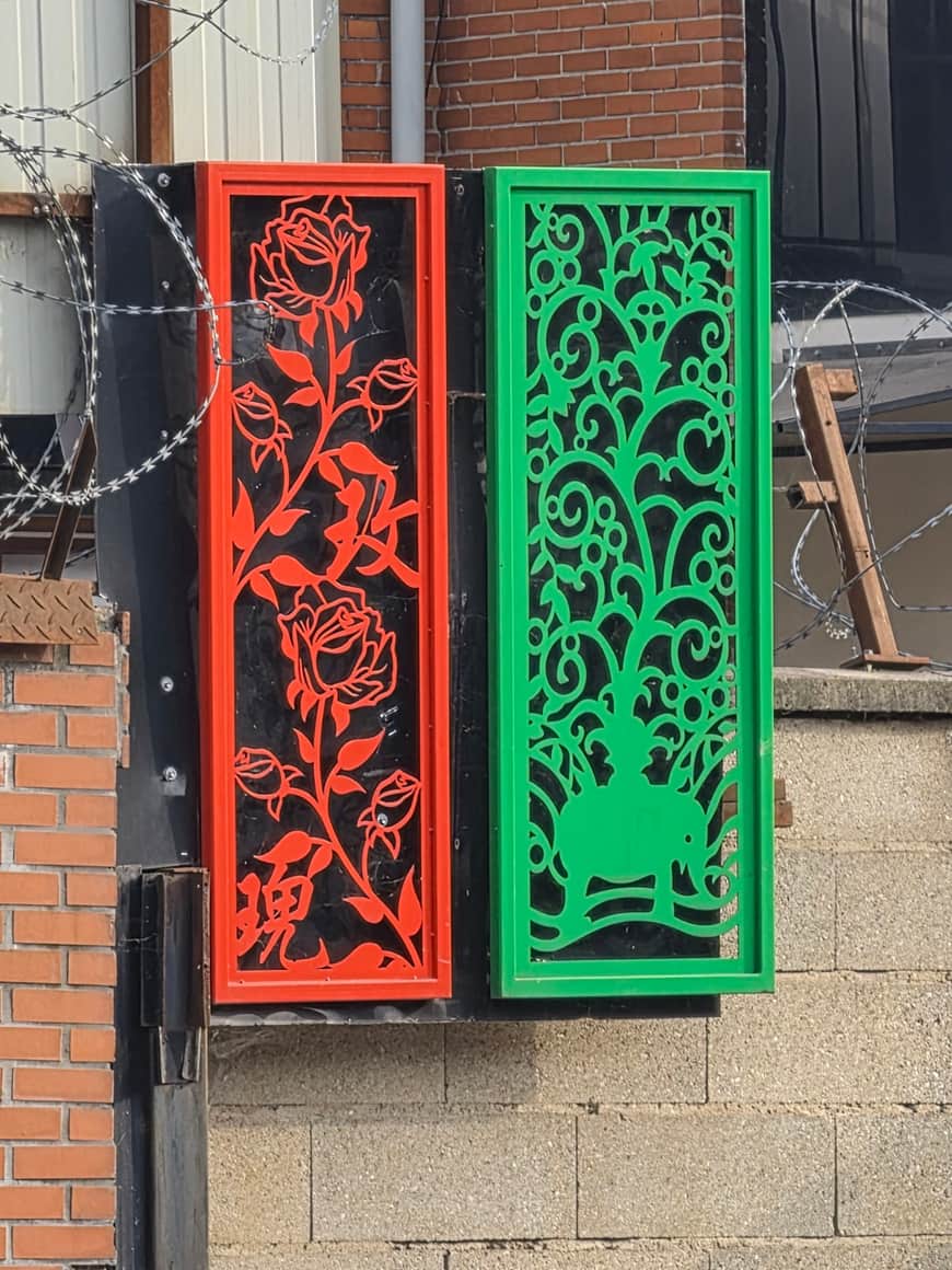 Deux panneaux métalliques ajourés rouge et vert, ornés de roses et d’un arbre, devant une clôture de barbelés