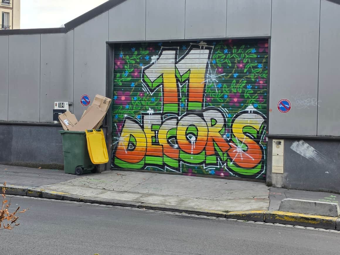 Rideau métallique peint d’un grand M et du mot Doors, entouré de fleurs, avec poubelles et cartons devant la façade