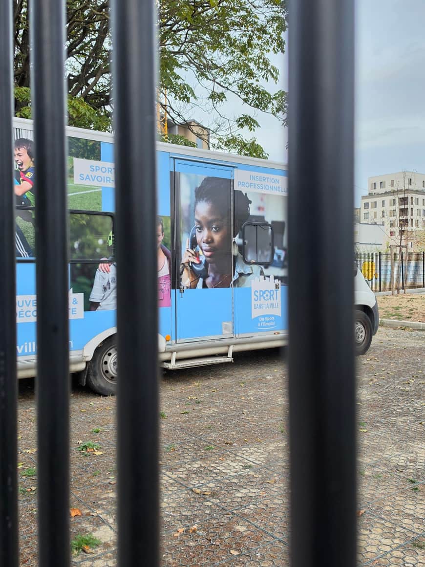 Camion consacré au sport et à l’insertion professionnelle photographié à travers les barreaux noirs d’une clôture