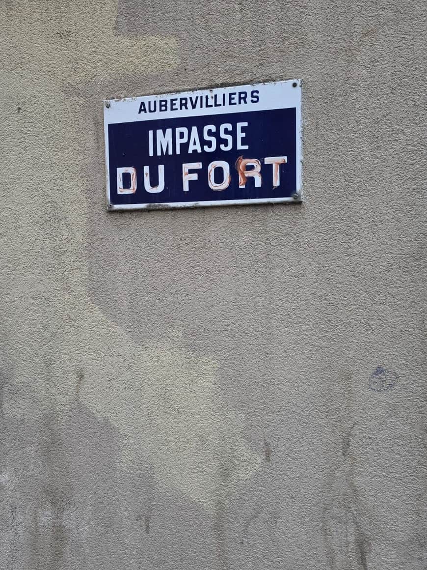 Plaque de rue bleue et blanche portant Aubervilliers, Impasse du Fort, sur un mur gris rugueux