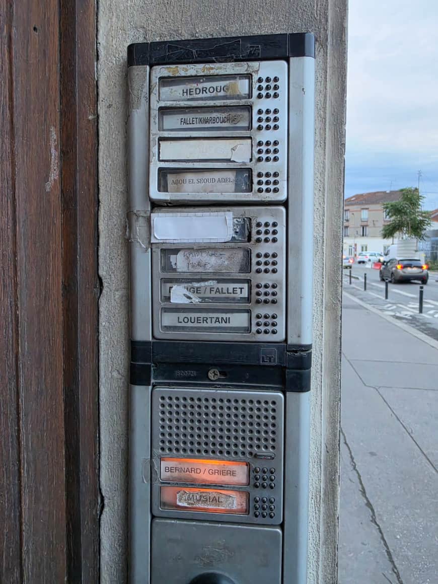 Interphone collectif ancien fixé entre une porte en bois et un mur, avec la rue visible en arrière-plan