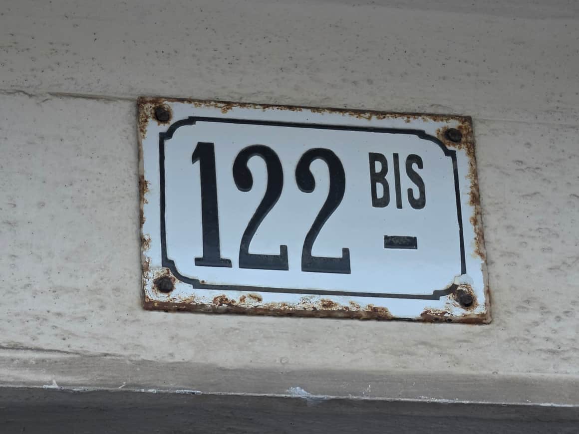 Plaque émaillée blanche à bord noir portant le numéro 122 bis, fixée par quatre vis rouillées