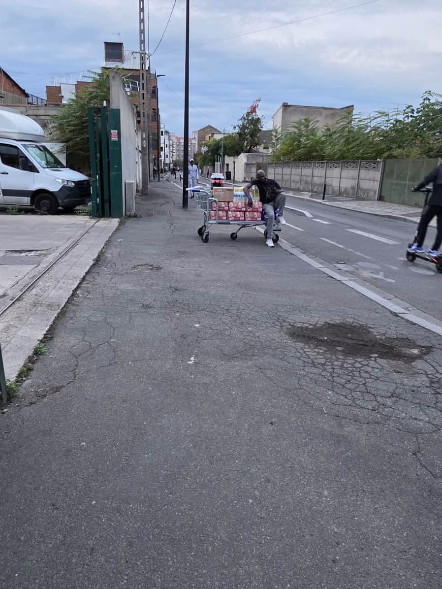 Vue large d’une rue où un homme pousse un chariot de supermarché chargé de cartons et de packs de boissons