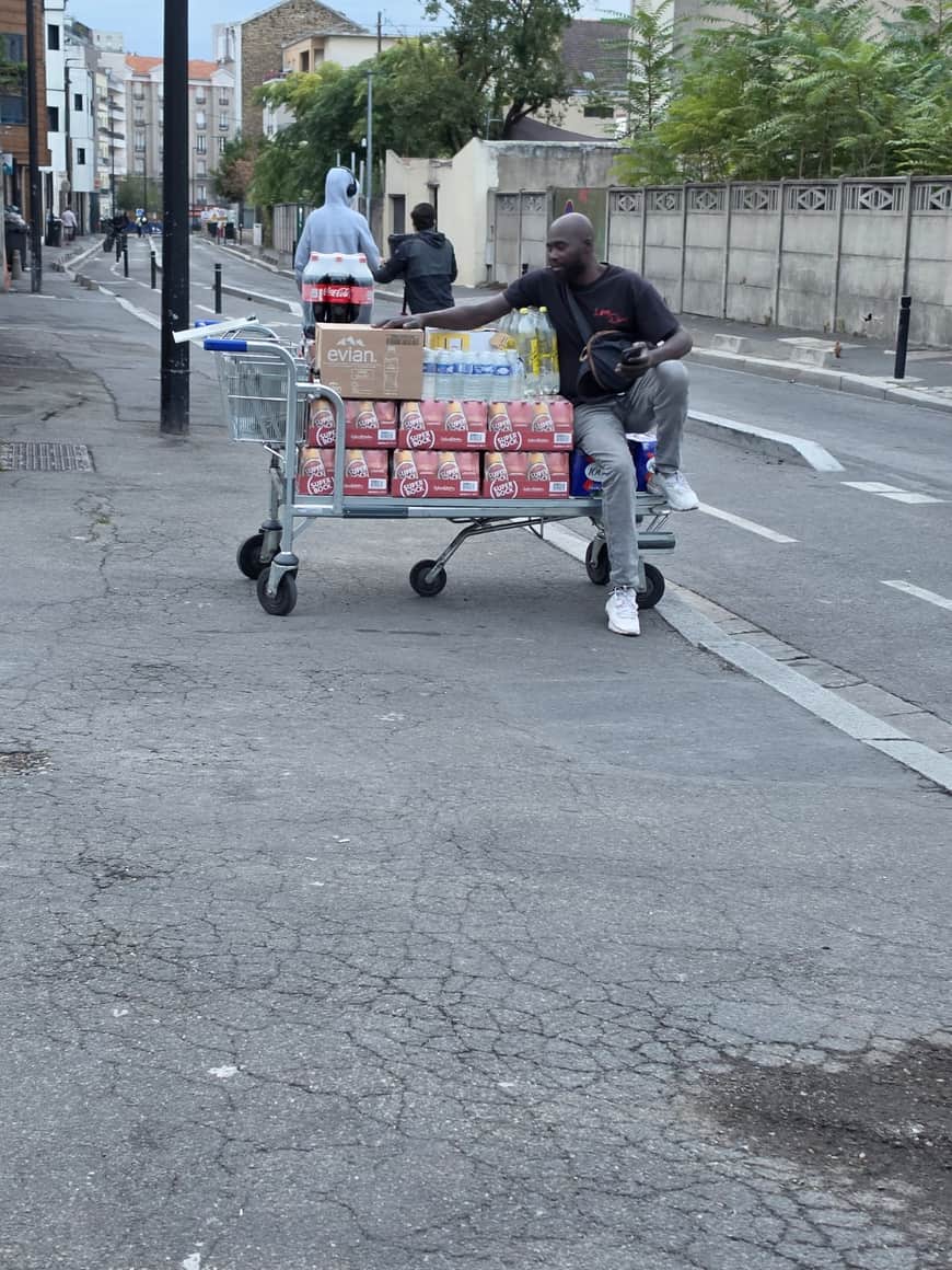 Homme poussant dans une rue un chariot de supermarché chargé de cartons et de packs de boissons