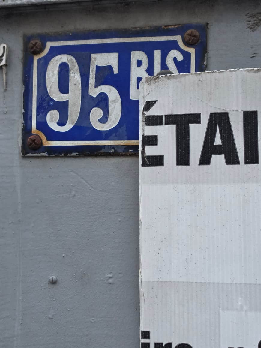 Plaque émaillée bleue portant le numéro 95 bis, partiellement masquée par un panneau de chantier blanc