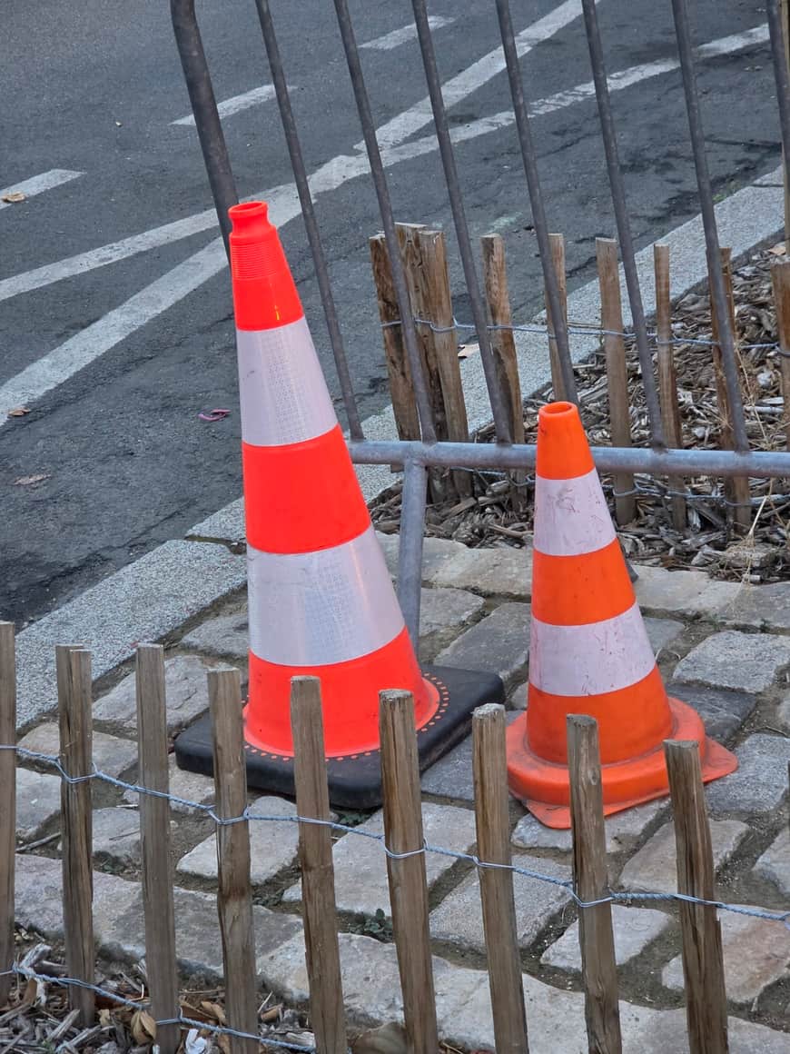 Deux cônes orange et blancs de tailles différentes disposés derrière une bordure basse en bois et une grille métallique
