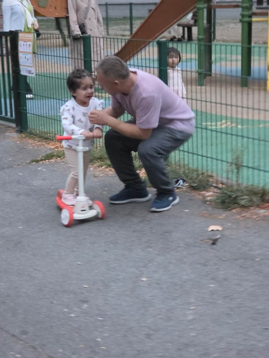 Jeune enfant en pleurs sur une trottinette et adulte penché vers lui, devant une aire de jeux clôturée