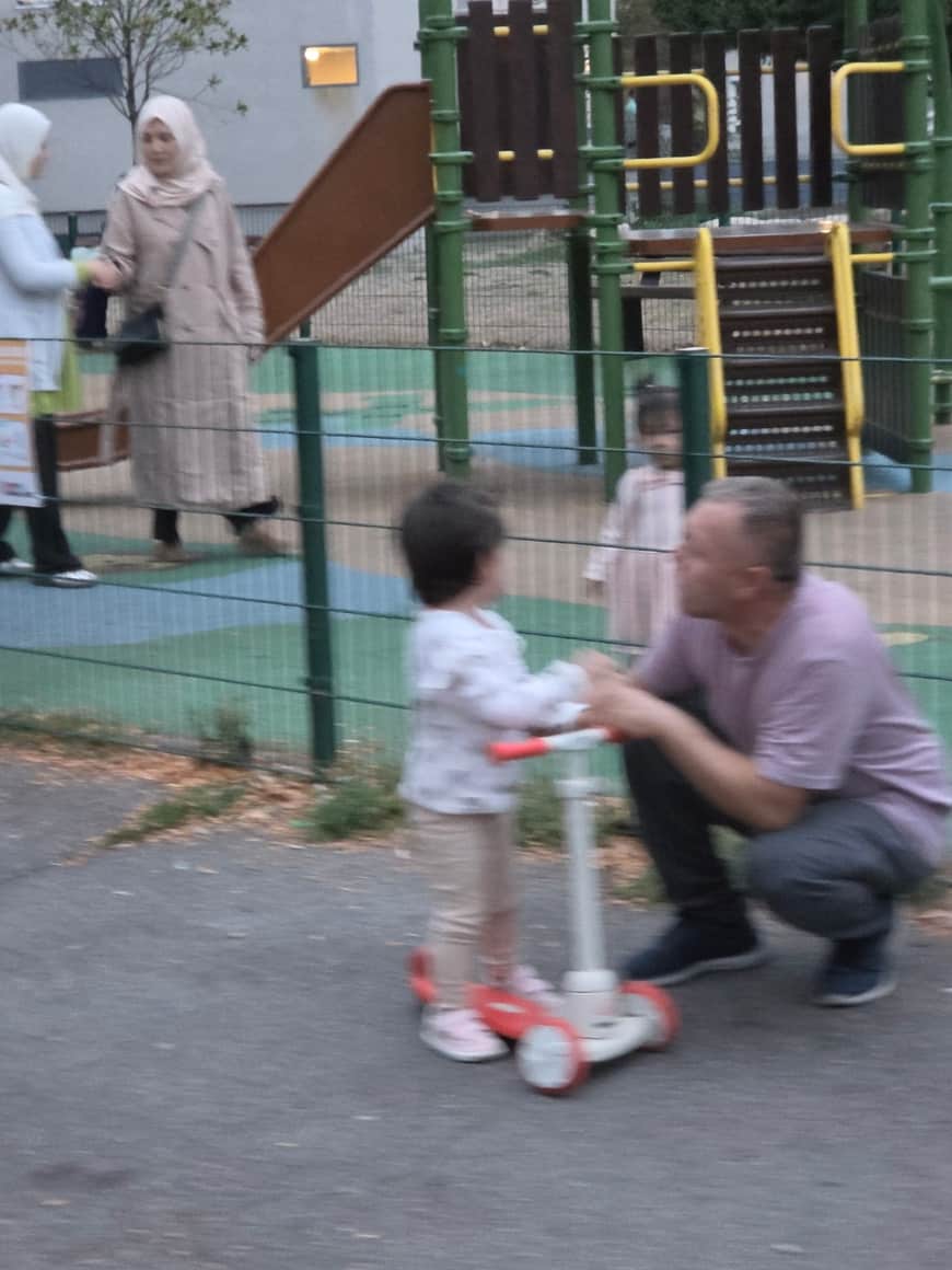 Adulte accroupi auprès d’un jeune enfant debout sur une trottinette, devant la clôture d’une aire de jeux