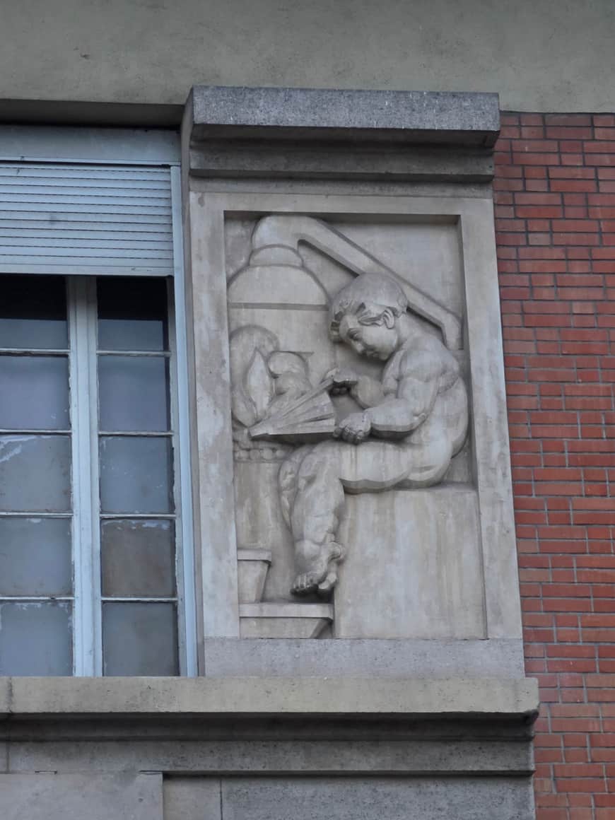 Relief de pierre représentant deux figures penchées sur un livre ouvert, entre une fenêtre et un mur de brique