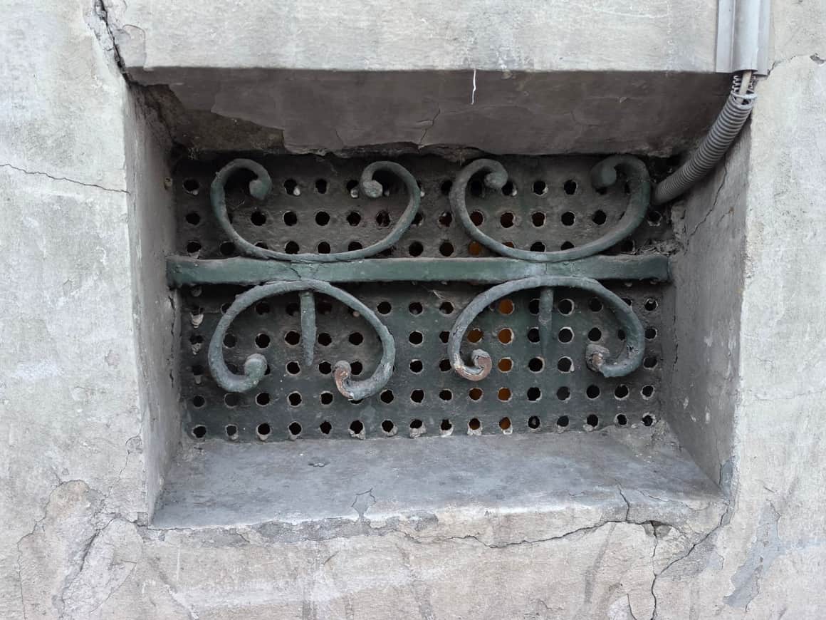 Ouverture basse dans un mur enduit, fermée par une plaque perforée et une ferronnerie verte à quatre volutes