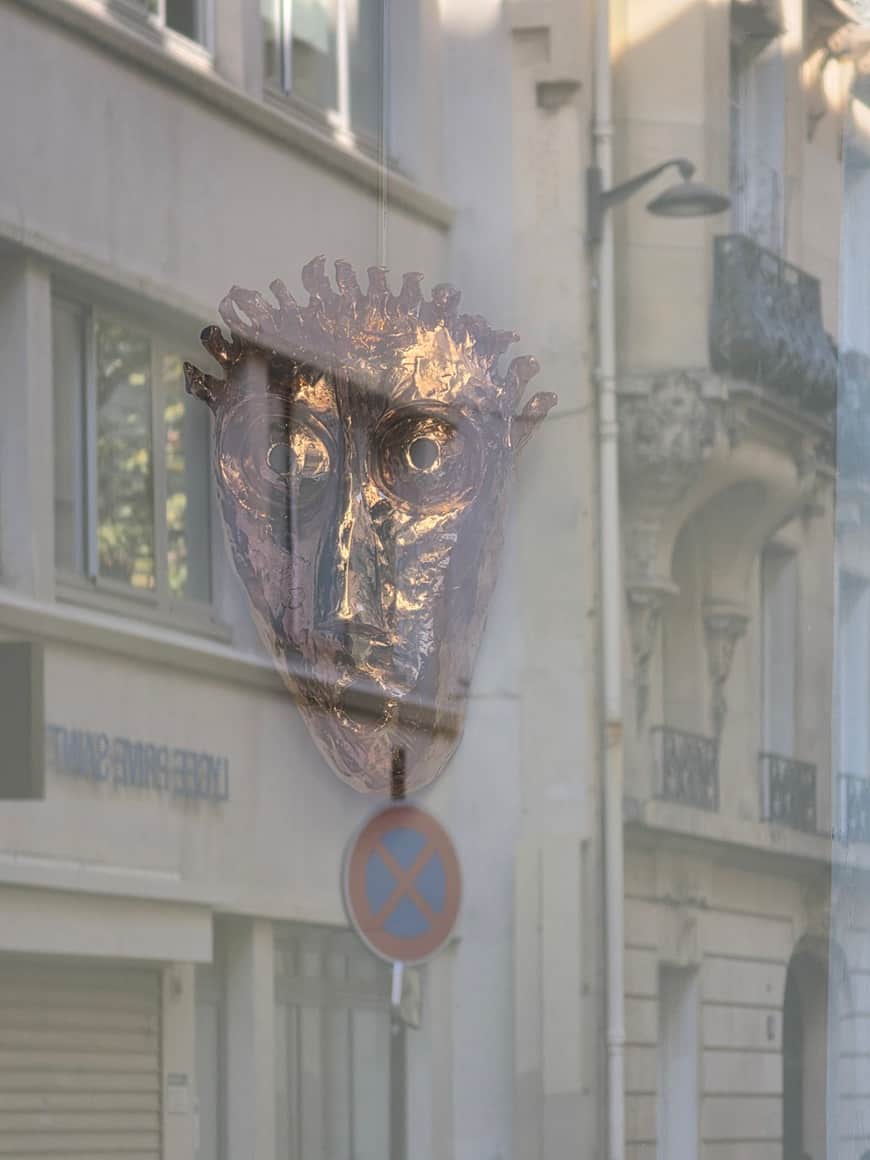 Masque métallique exposé derrière une vitre où se reflètent une rue et un panneau de stationnement