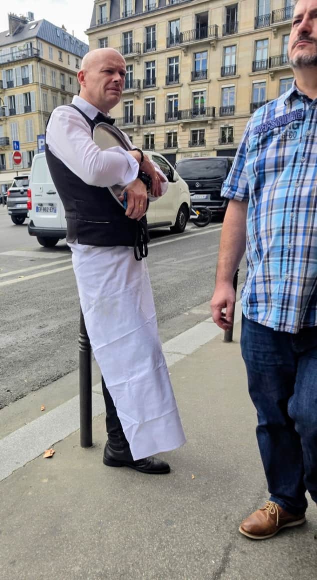 Serveur en tablier blanc tenant un plateau sous le bras auprès d’un passant en chemise à carreaux