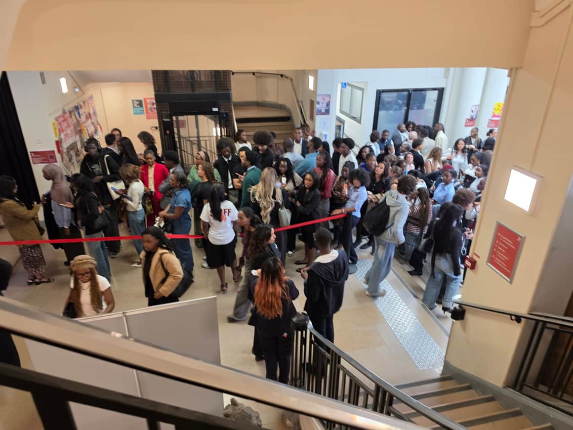 Foule réunie dans un hall entre un escalier, un ascenseur et un ruban rouge