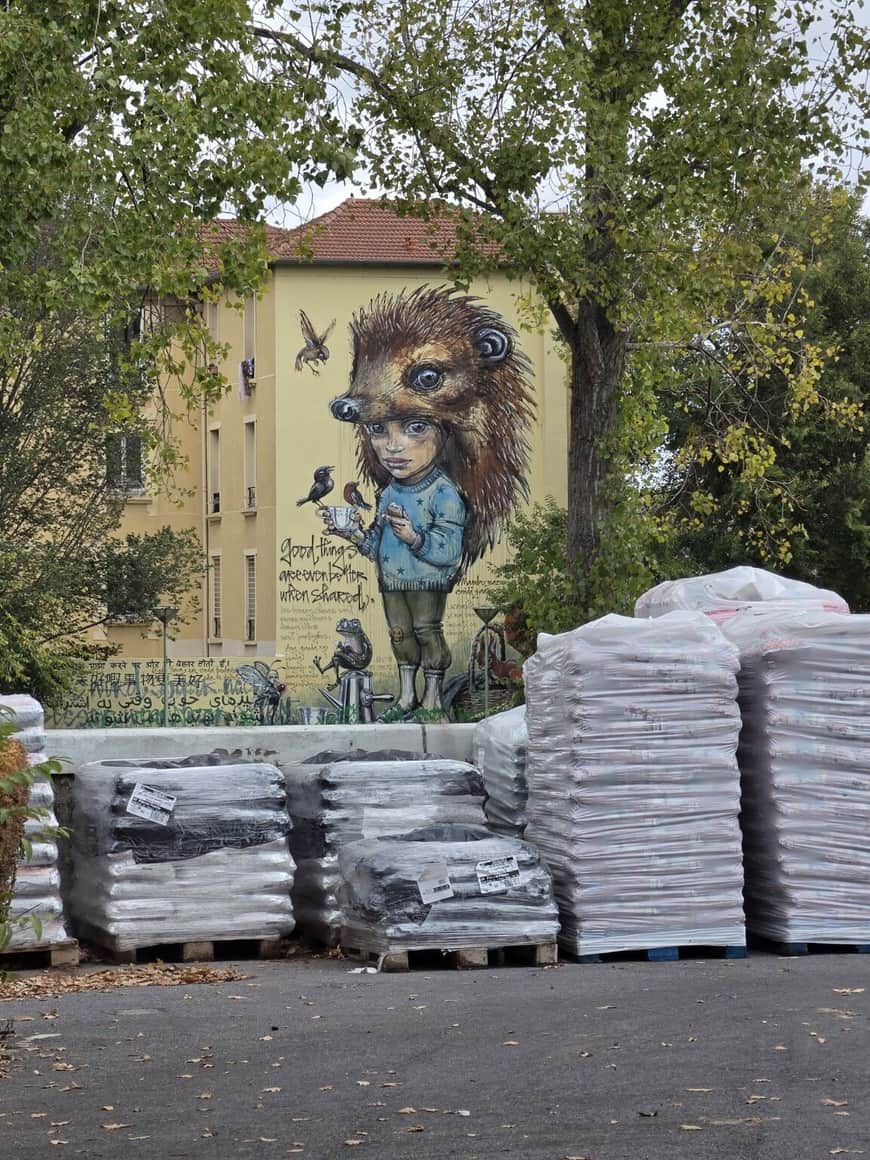 Peinture murale d’une enfant coiffée d’un immense hérisson, derrière des palettes de matériaux emballés et parmi les arbres