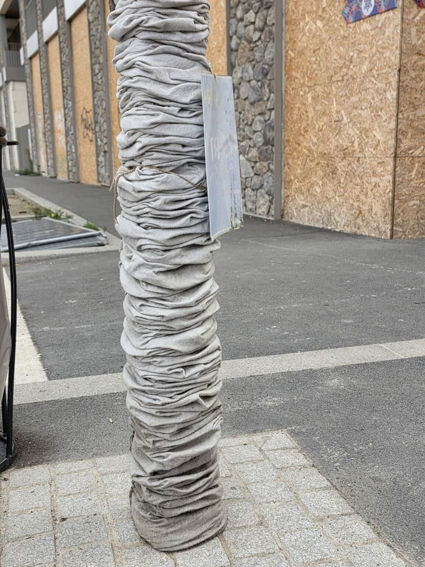 Poteau entièrement enveloppé d’un textile gris plissé et serré par une corde devant une façade obturée