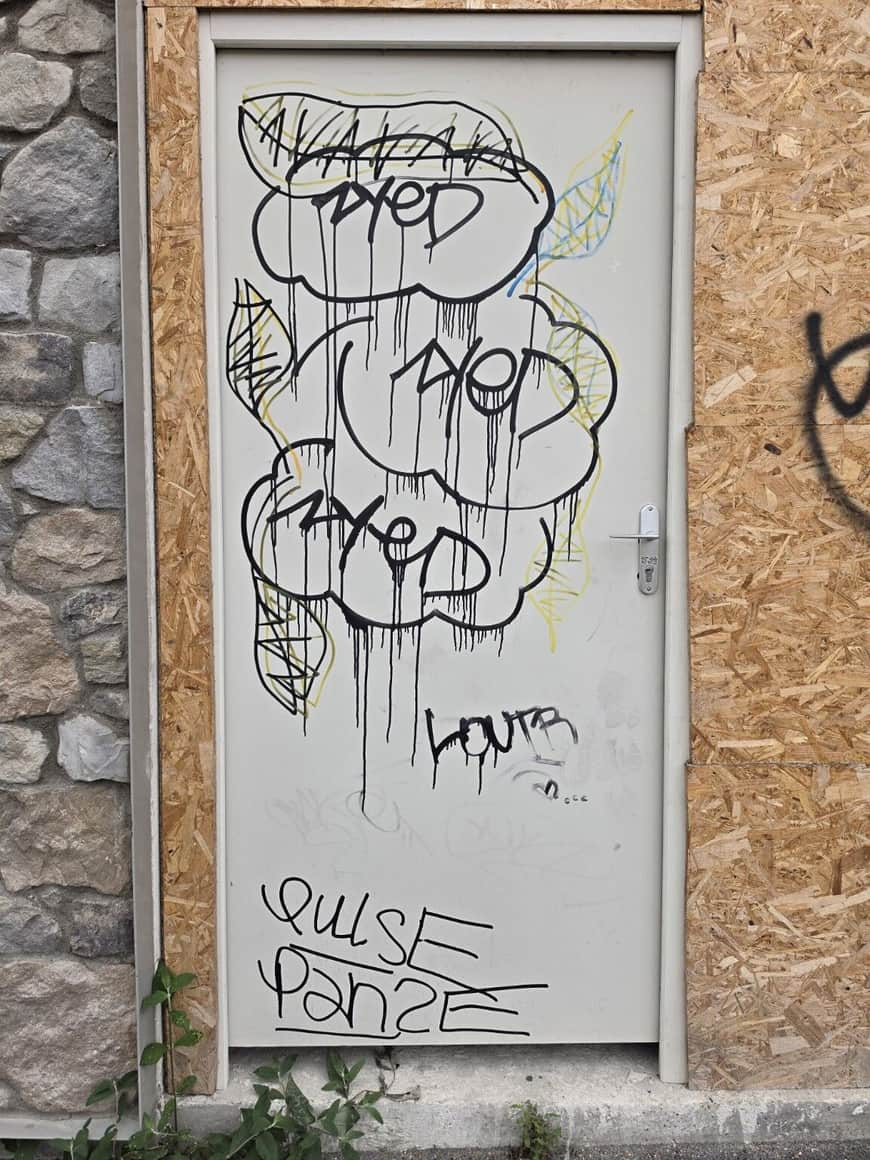 Porte blanche couverte de graffitis noirs, jaunes et bleus, encadrée de pierre et de panneaux de bois aggloméré