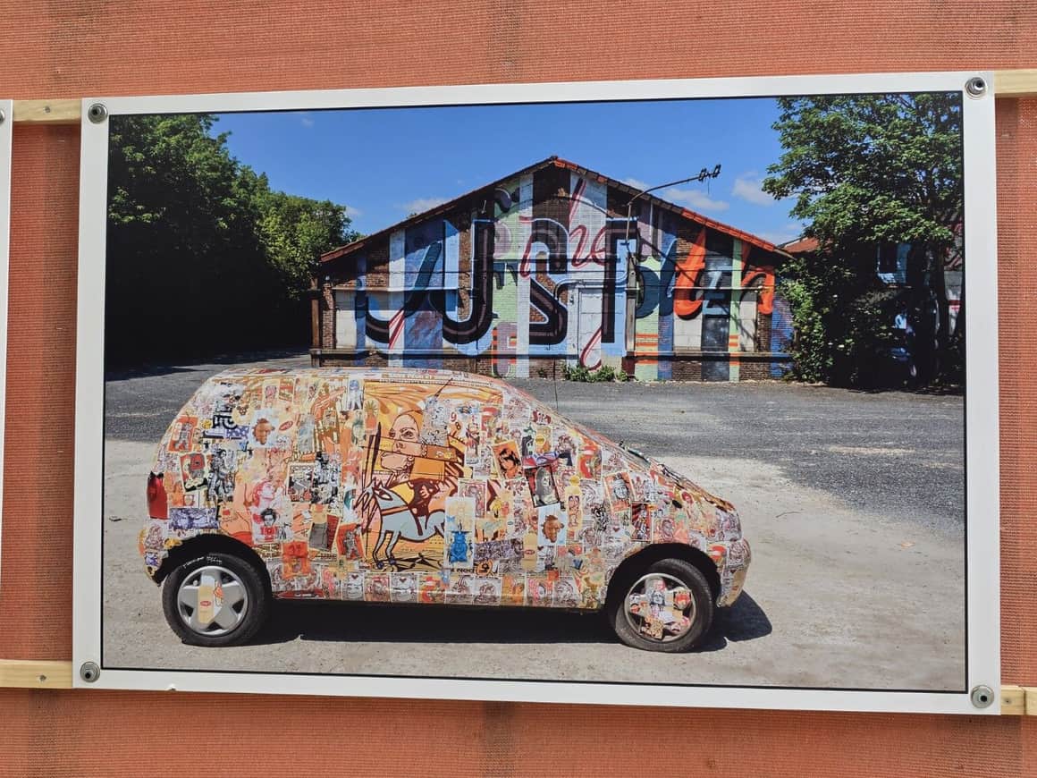 Photographie fixée sur une toile orange montrant une petite voiture couverte d’autocollants devant un bâtiment peint