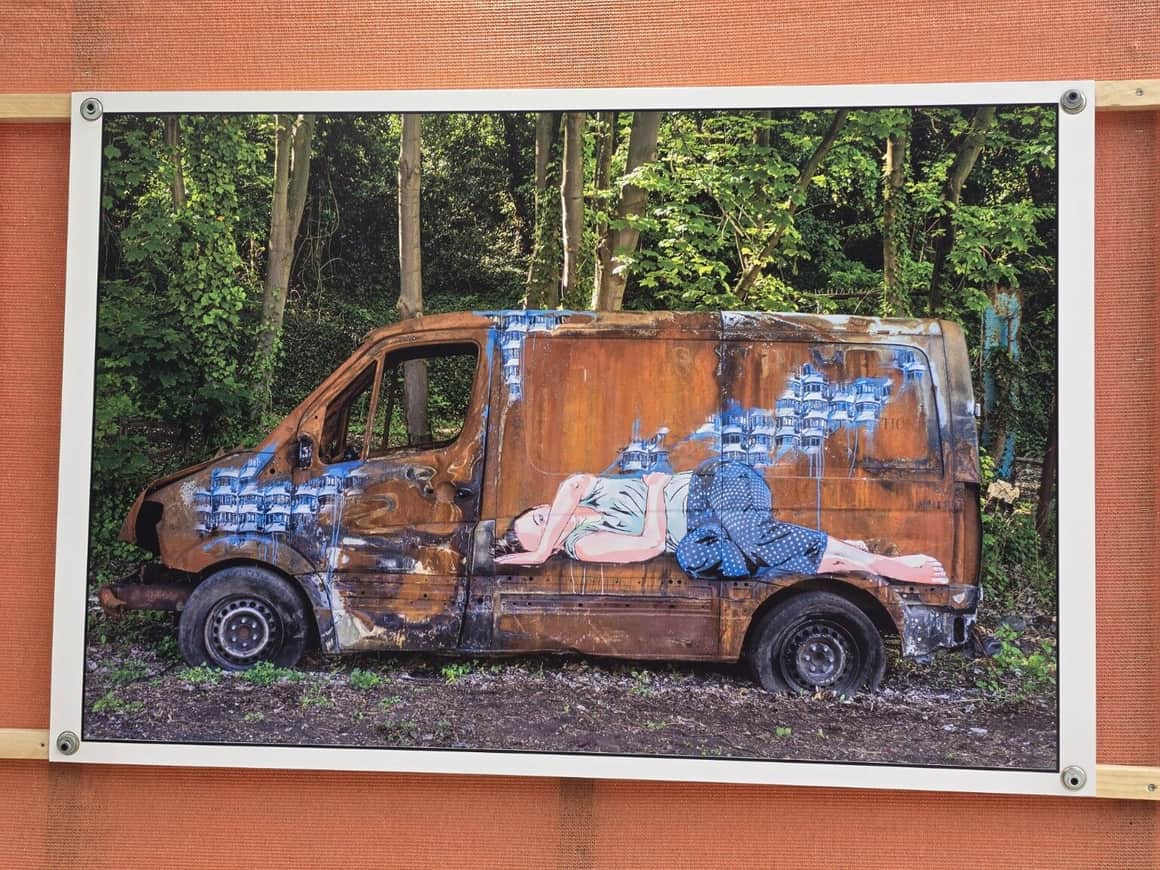 Photographie fixée sur une toile orange montrant une camionnette rouillée peinte d’une figure féminine dans un terrain boisé