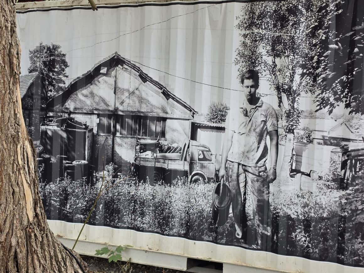 Photographie noir et blanc d’une cour industrielle avec camion et personnage, fixée sur une palissade orange
