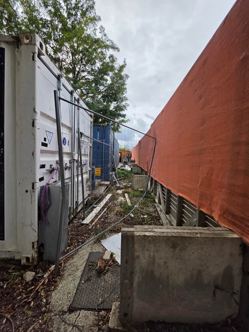 Passage de chantier entre conteneurs blancs et bleus, clôture grillagée et écran orange