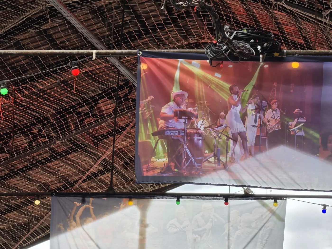 Toile montrant un spectacle, suspendue sous un plafond grillagé et photographiée avec son environnement