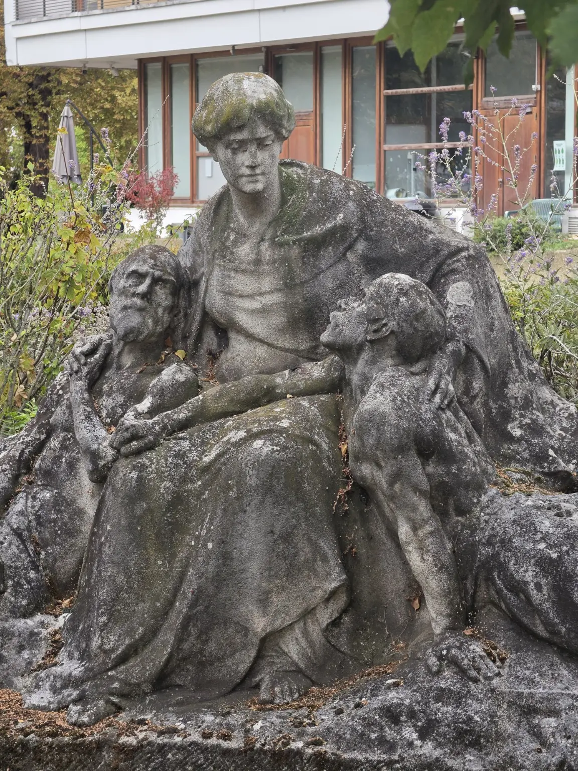 Groupe sculpté en pierre patinée représentant une figure assise entourée de deux figures plus petites, dans un jardin devant un bâtiment moderne
