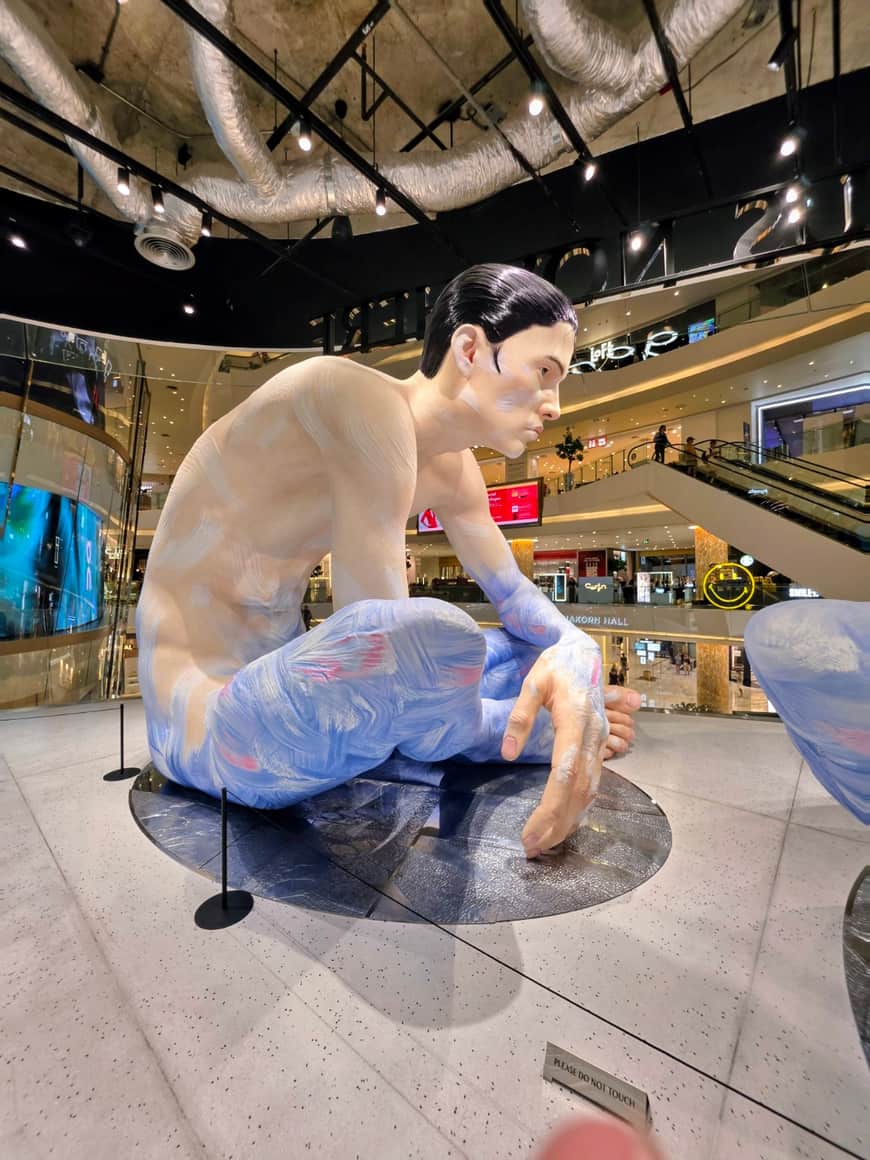 Sculpture figurative accroupie au centre d’un espace commercial.