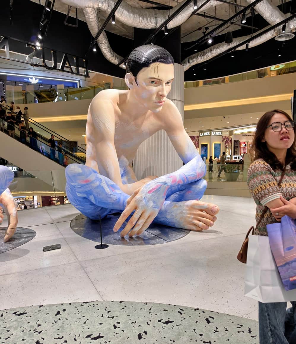 Sculpture figurative accroupie dans une galerie commerciale, avec des visiteurs à l’arrière-plan.