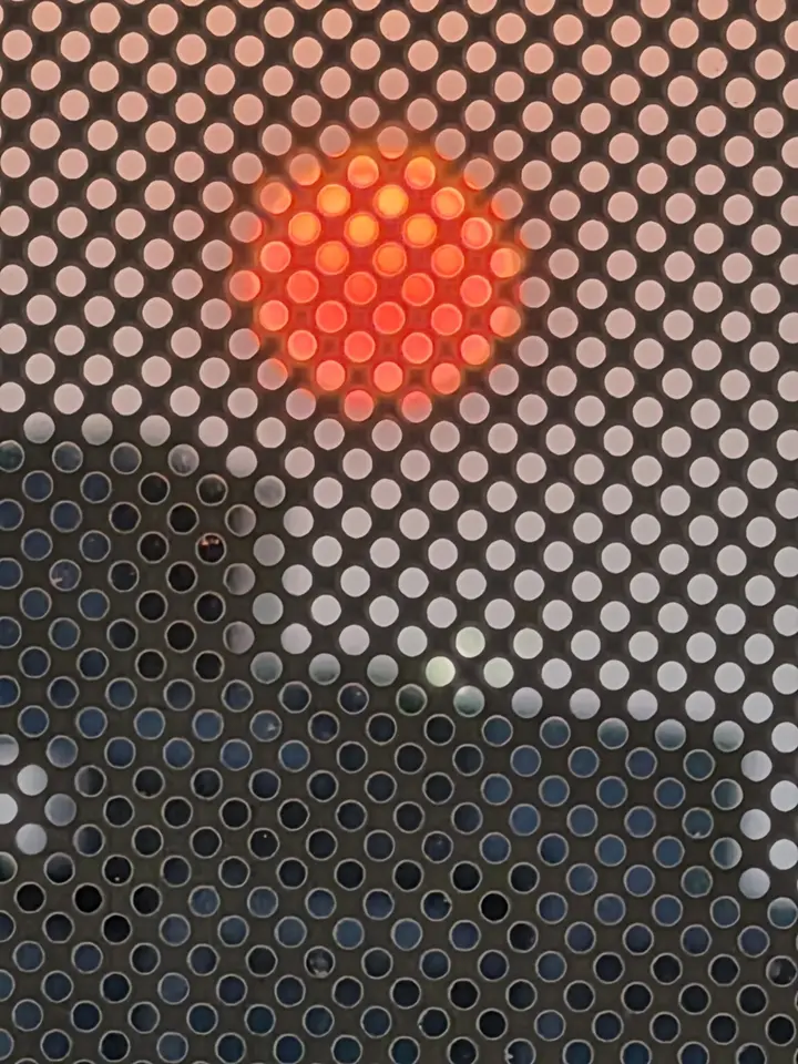 Disque solaire rouge vu à travers une grille métallique perforée.