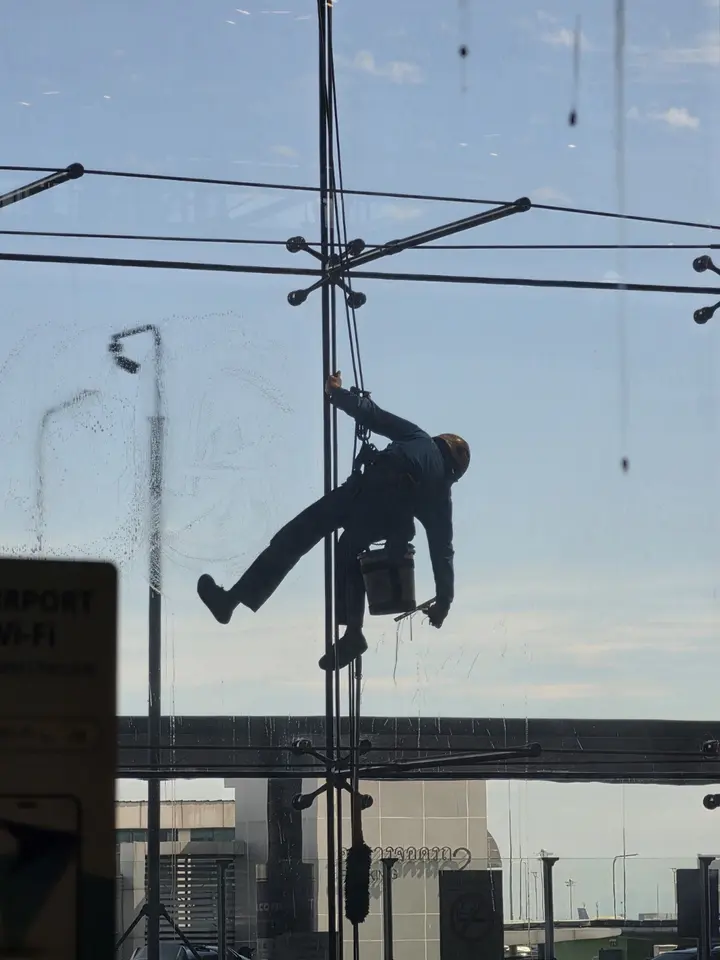 Laveur de vitres suspendu par des cordes devant une vaste façade de verre et un ciel bleu.