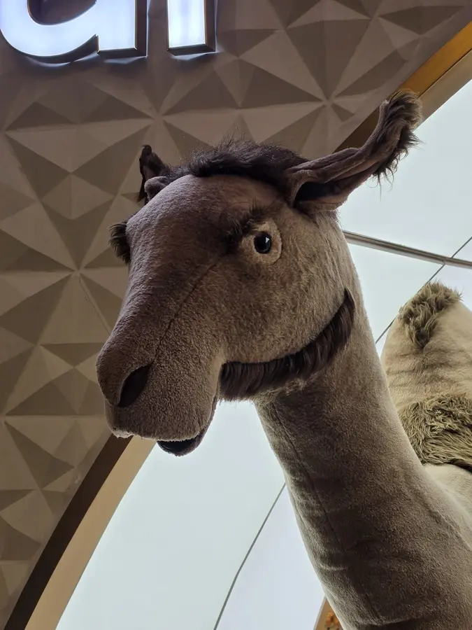 Grand animal de peluche au long cou, photographié en contre-plongée sous un plafond géométrique.