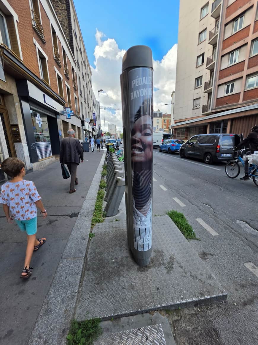 Colonne cylindrique portant les mots Pédalez, rayonnez, au bord d’une piste cyclable dans une rue urbaine.